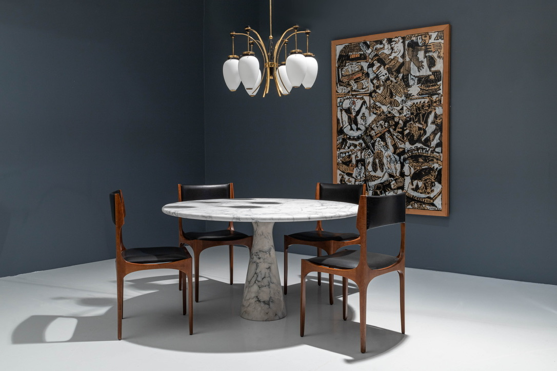 Round marble dining table with four leather and wood dining chairs, a brass and opaline chandelier and a large framed artwork on a dark blue wall